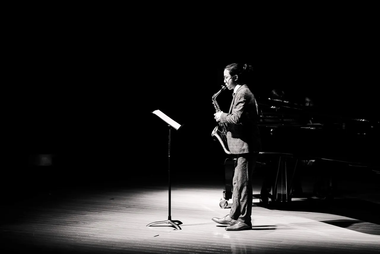 Aaron playing alto saxophone in a single spotlight on a dark concert stage, a music stand beside him and a grand piano behind.
