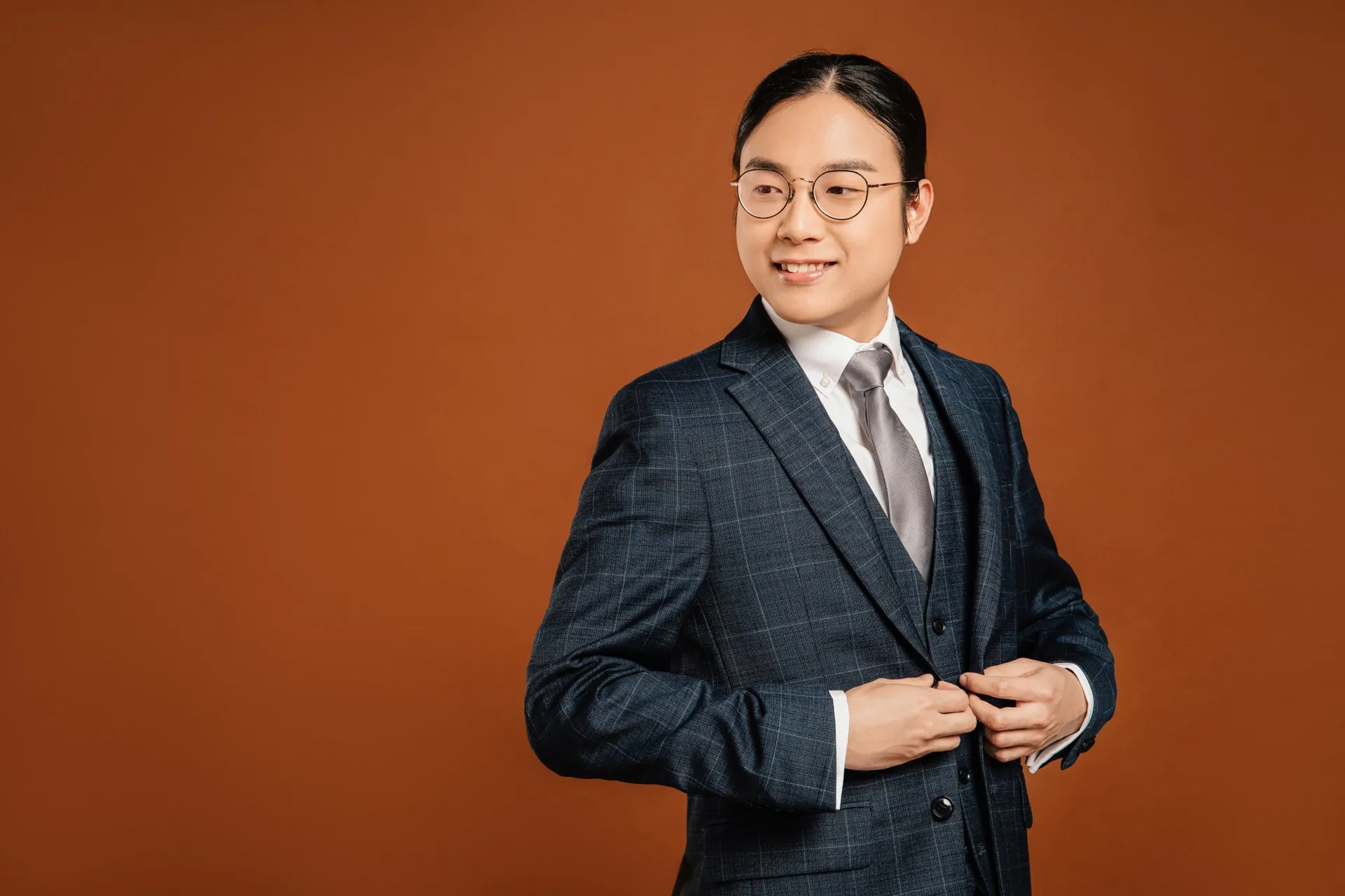Portrait of Aaron smiling and looking over his shoulder, in a navy checked suit against an orange backdrop.