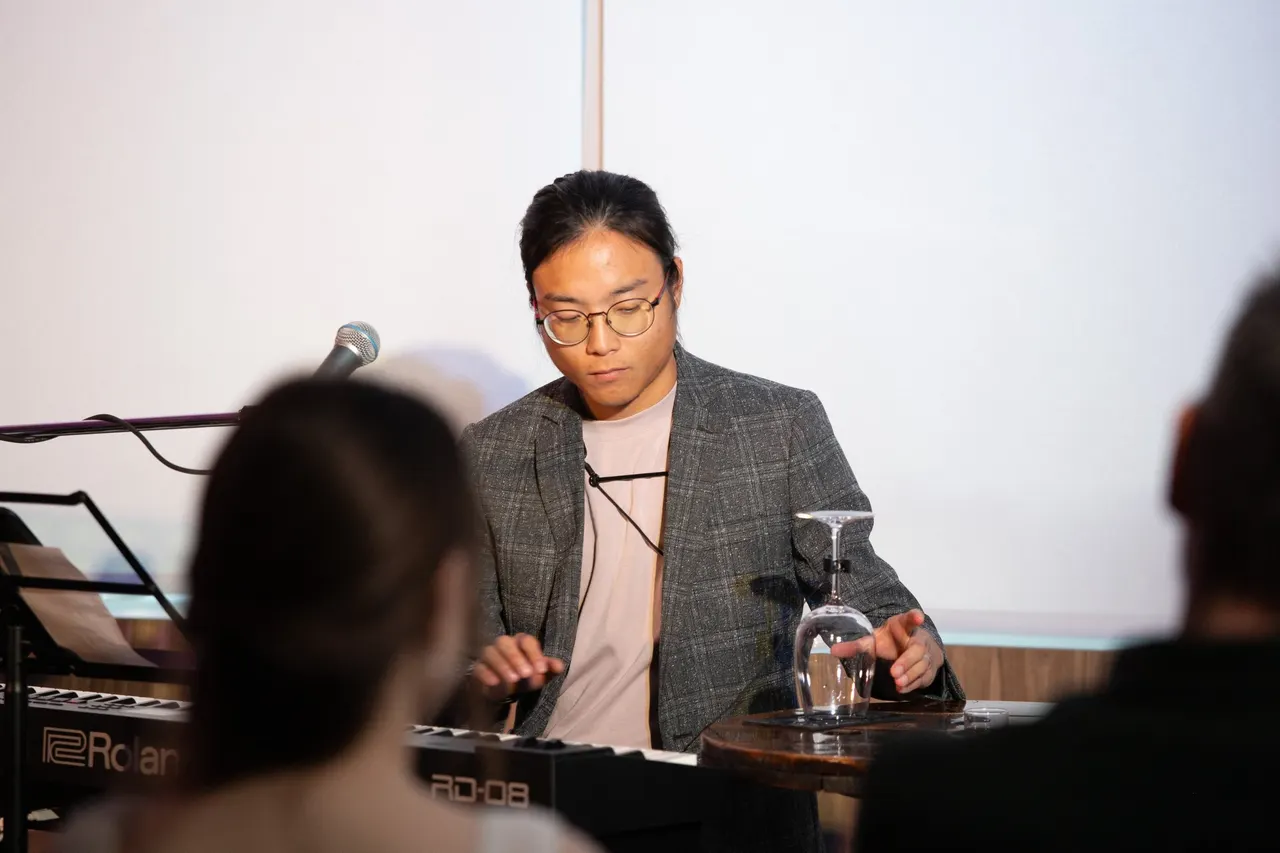 Aaron looking down at a keyboard with both hands raised above it, an upturned wine glass on the table beside him and the heads of the audience in the foreground.