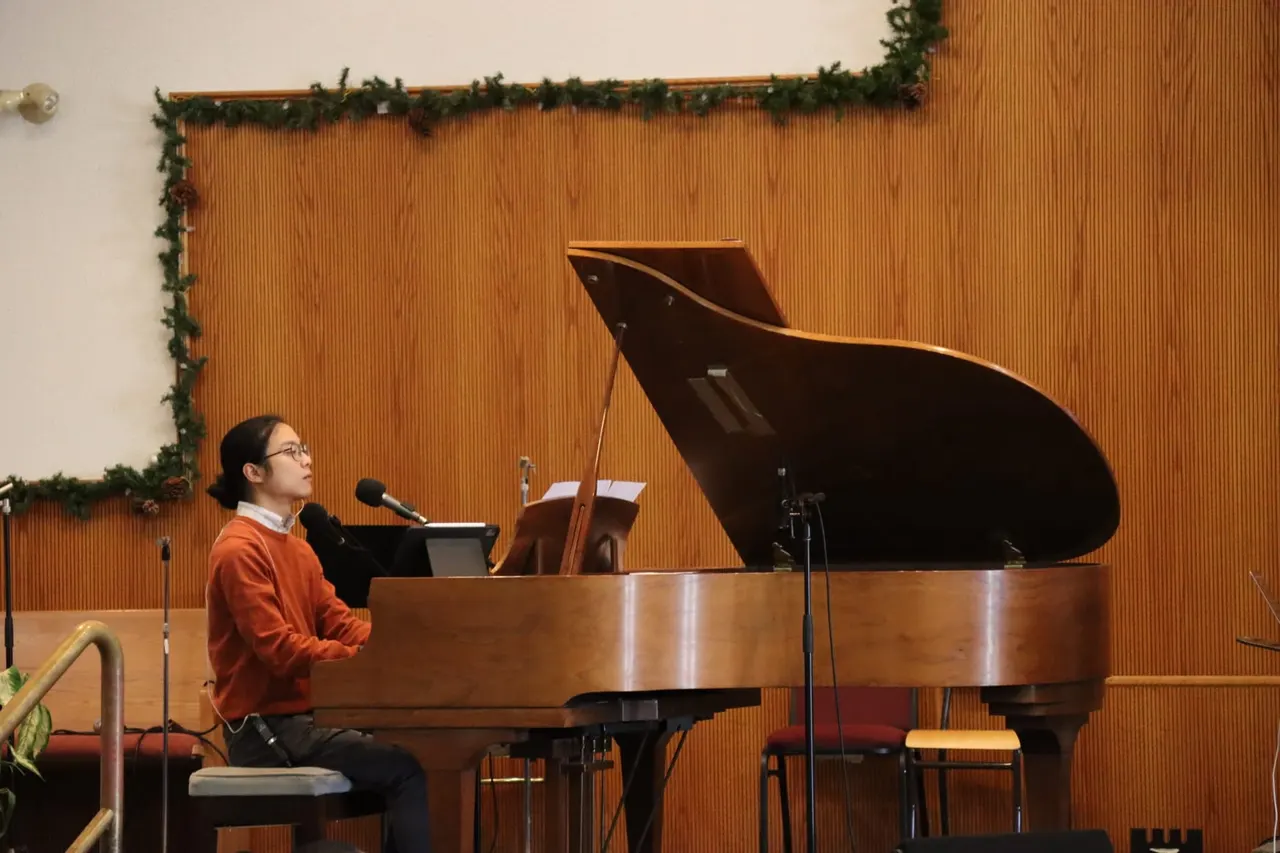 Aaron in a rust-coloured sweater playing a grand piano in front of a wooden wall hung with evergreen garland.