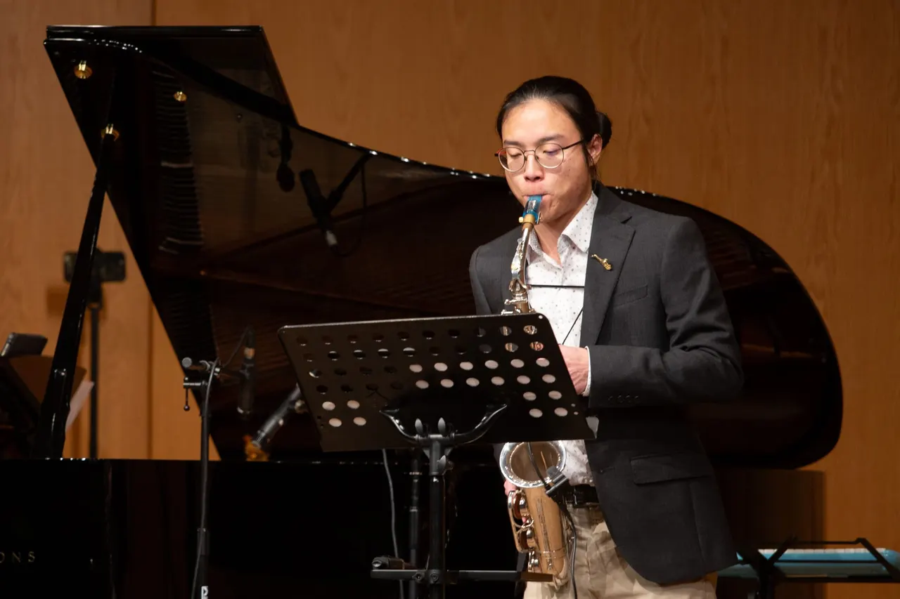 Aaron playing alto saxophone from a music stand in a concert hall, a grand piano and warm wooden wall behind him.