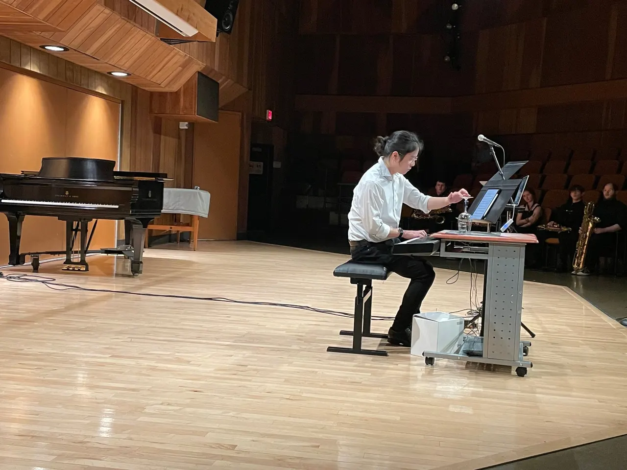 Aaron seated at a keyboard on a wide wooden stage, reaching toward an upturned wine glass, with a grand piano to one side and listeners in the dark seats behind.